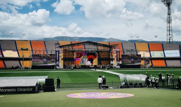 A sports stadium with a large stage set up on the field, hosting an outdoor event. Spectators wearing colorful hats sit in the foreground. The seating areas display vibrant colors, and a clear blue sky with scattered clouds is visible. Large screens display graphics related to the event.