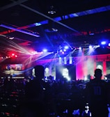 A dimly lit indoor arena filled with an audience. Bright stage lights in red and blue illuminate multiple large screens displaying graphics.