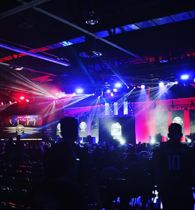A dimly lit indoor arena filled with an audience. Bright stage lights in red and blue illuminate multiple large screens displaying graphics.