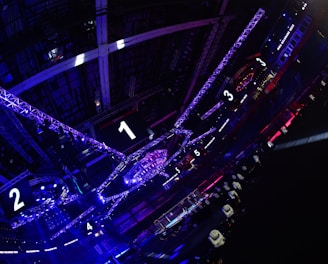 A large indoor arena with colorful and vibrant lighting, featuring a center stage surrounded by rigging and structures. Large numbered screens are visible, indicating marked sections from one to five. Various pieces of equipment and people are scattered around the venue.