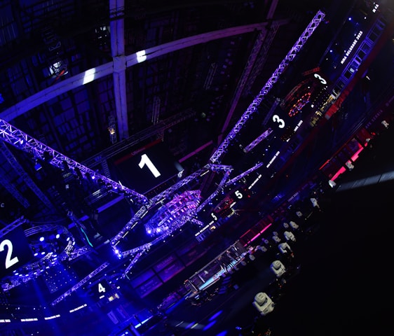 A large indoor arena with colorful and vibrant lighting, featuring a center stage surrounded by rigging and structures. Large numbered screens are visible, indicating marked sections from one to five. Various pieces of equipment and people are scattered around the venue.