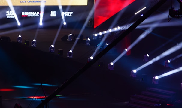 An array of bright stage lights beams across a dark arena, creating a dynamic atmosphere with a large screen displaying various logos and text. A camera crane is visible in the foreground, suggesting a professional event setup.