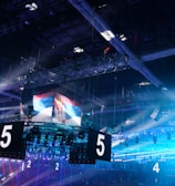 A large indoor arena is equipped with advanced lighting and audio systems. Suspended above the space are numerous digital displays showcasing numbers. The environment is filled with blue illumination and spotlights creating a vibrant atmosphere.