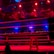 An empty boxing ring is illuminated by dramatic red and blue spotlights inside a dimly-lit arena. A small stool is placed in the center of the ring, surrounded by ropes. The arena is adorned with banners depicting action scenes, and a few silhouettes of spectators can be seen in the background.
