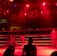 A wrestling ring is illuminated with red lighting, creating a dramatic atmosphere. Several people are visible around the ring, with silhouetted spectators in the foreground. Banners or art adorn the upper walls, and the venue has an industrial ceiling with exposed beams.