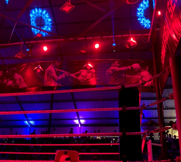 A brightly lit boxing or wrestling ring with vivid red and blue lights. Banners with images of fighters hang from the ceiling. The ropes of the ring are prominently visible, and a stool is placed inside the ring. The arena is filled with an audience, silhouetted against the colorful lighting.
