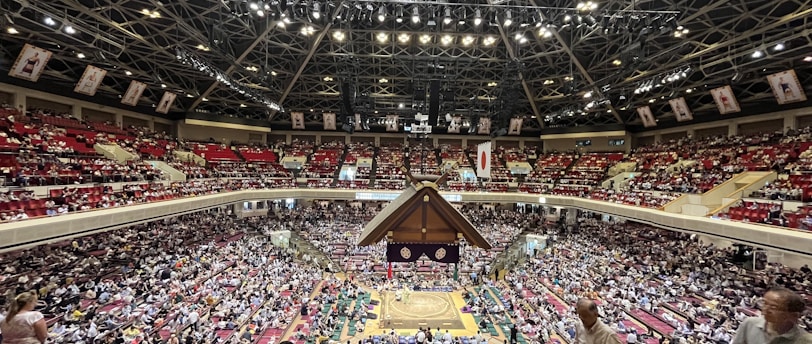 A large indoor arena packed with spectators seated in tiers around a central platform. The platform features traditional Japanese decor, indicating it may be used for sumo wrestling. The atmosphere is bustling, with people filling the seats and many spectators visible in the foreground.